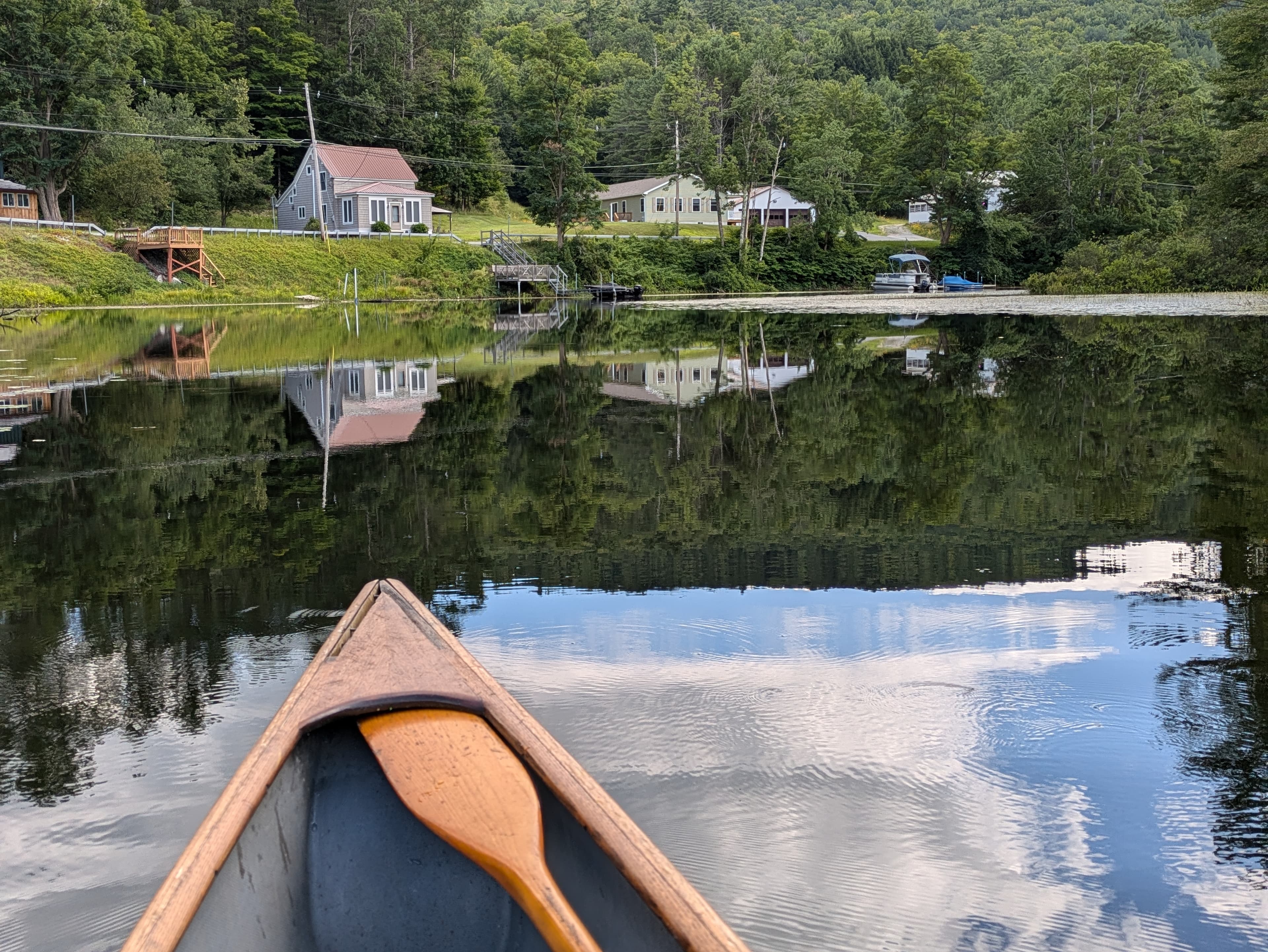 The bow of a canoe points across calm Brant Lake towards reflected lakeside buildings and densely wooded hills.